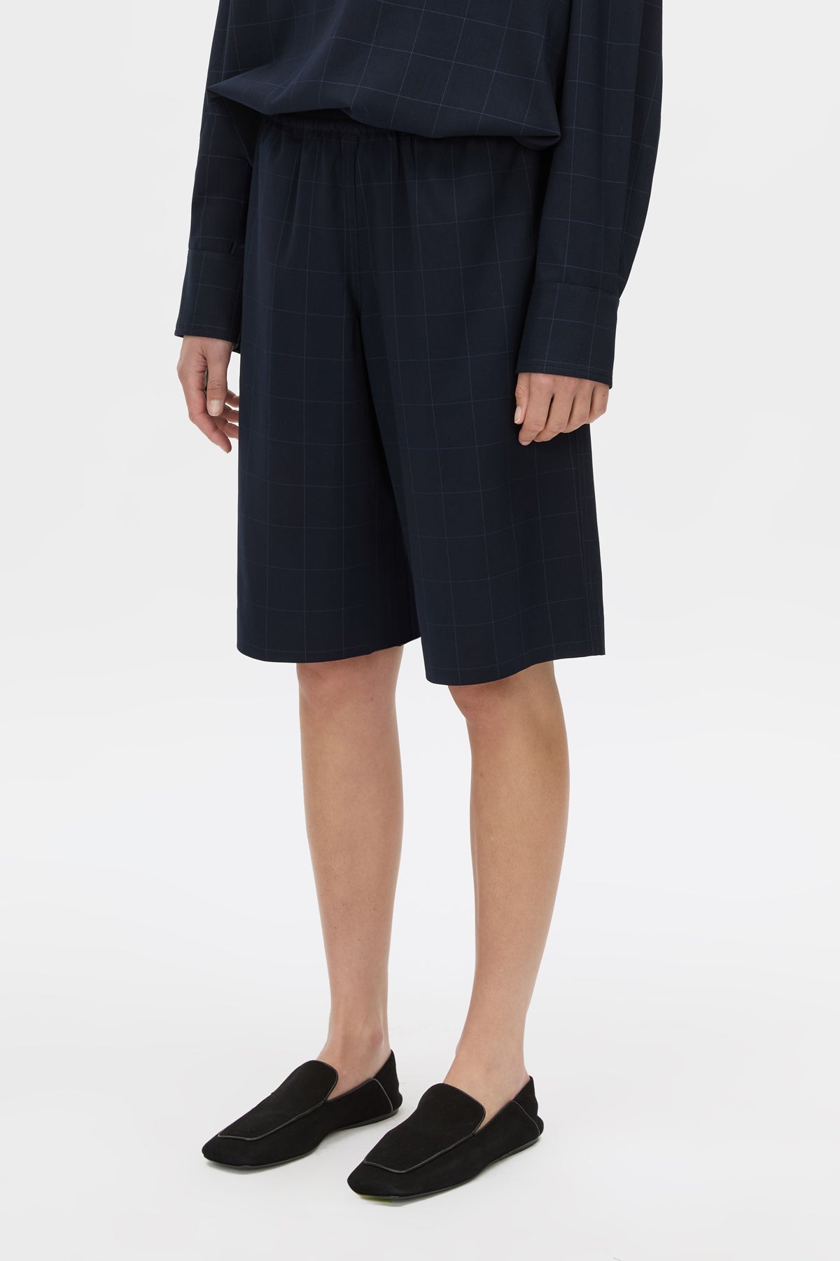 A model wearing dark navy blue plaid shorts, a matching navy top, and black slip-on loafers, shown from the waist down on a light gray studio background.
