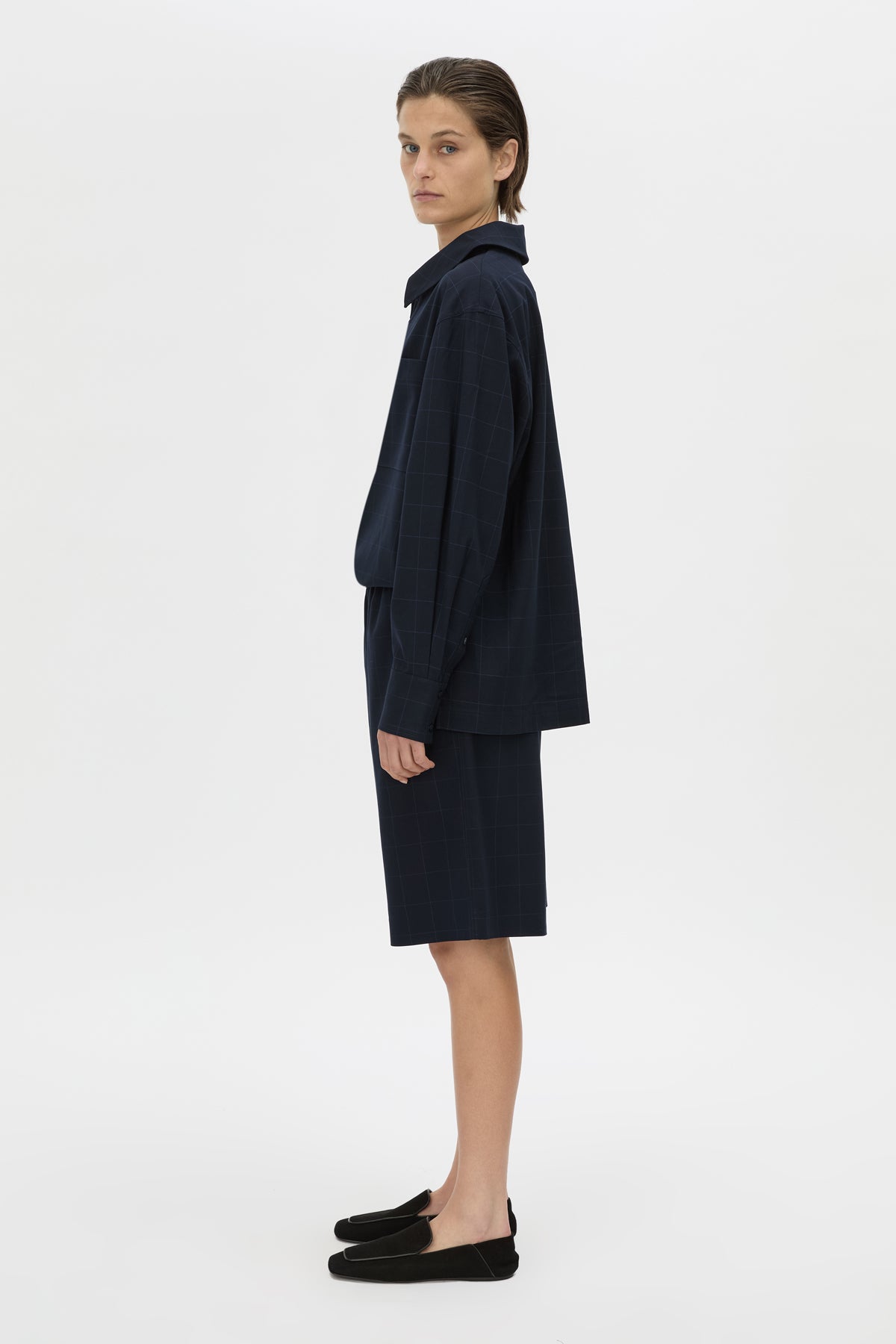 A model is shown in profile wearing a dark navy blue two-piece outfit consisting of a long-sleeve collared jacket and a matching knee-length skirt, paired with black slip-on loafers, against a plain light gray studio background.