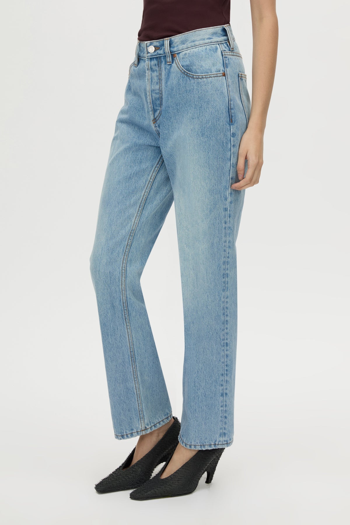 A model wearing light-wash blue denim jeans with a straight-leg or wide-leg fit, paired with black pointed-toe heels against a plain light gray studio background.