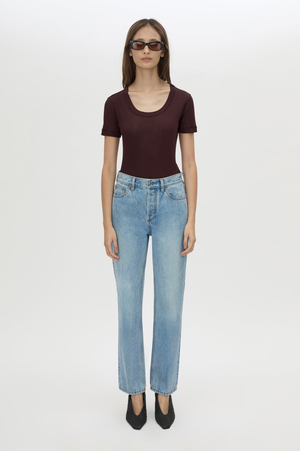 A model stands against a light gray studio background wearing a dark burgundy short-sleeve scoop-neck top, light-wash blue jeans, and black pointed shoes.