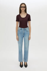 A model stands against a light gray studio background wearing a dark burgundy short-sleeve scoop-neck top, light-wash blue jeans, and black pointed shoes.