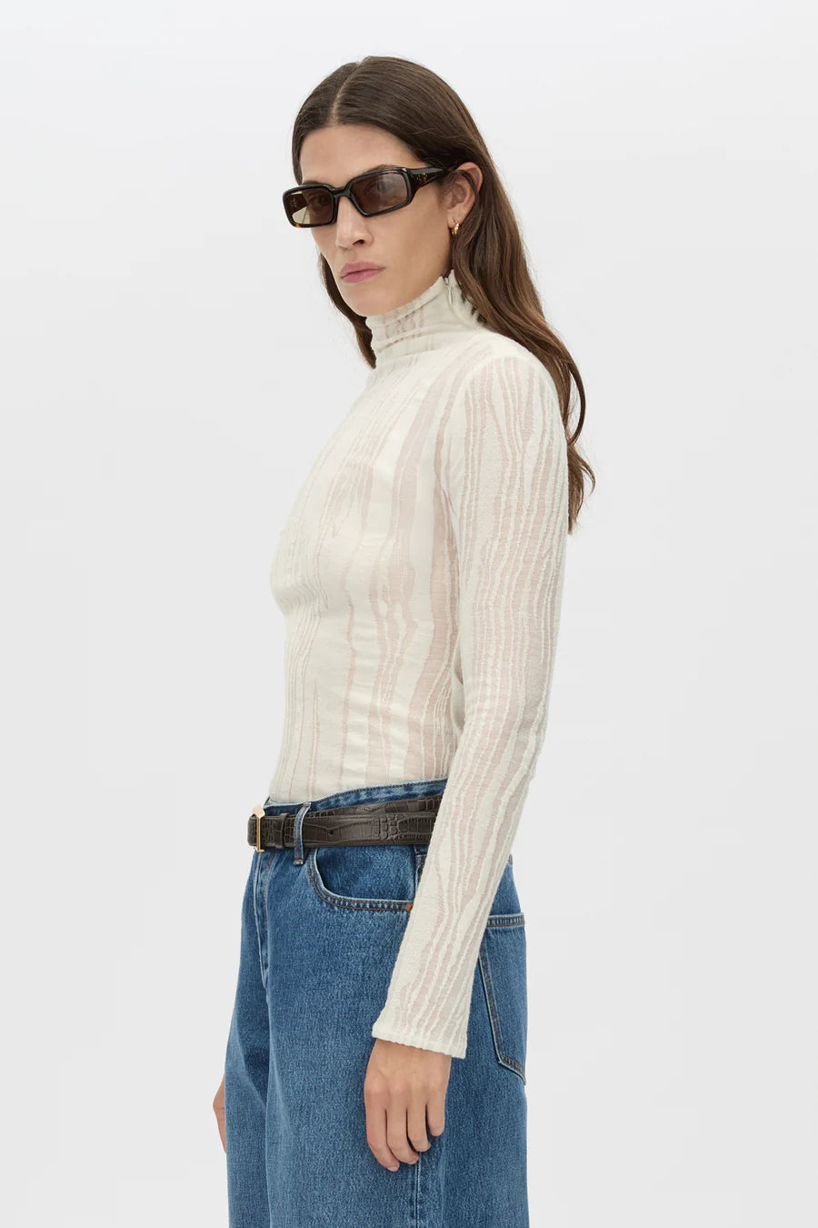 Woman wearing a cream ribbed turtleneck sweater and blue jeans on a white background