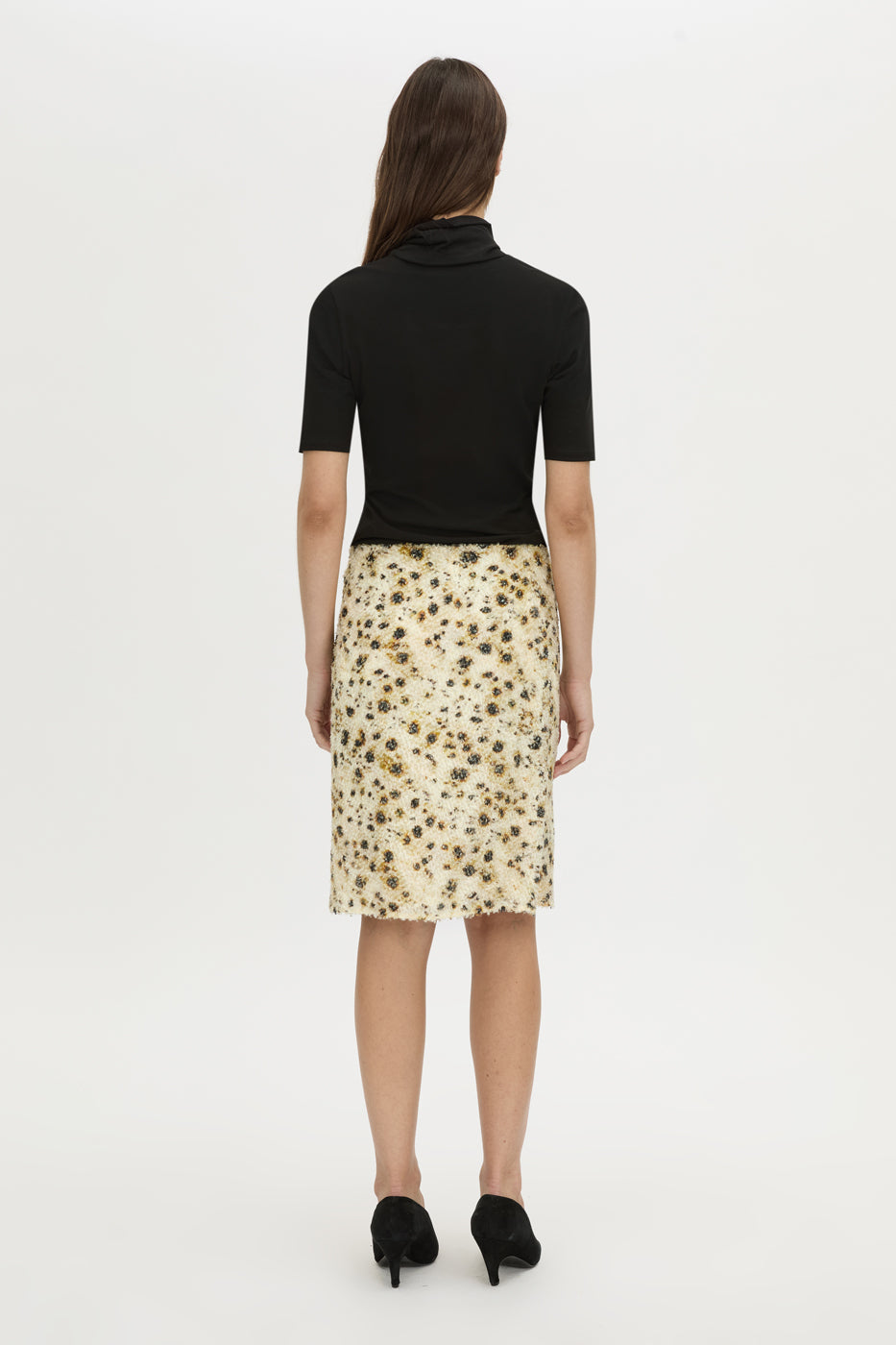 A woman shown from the back wearing a black short-sleeve mock-neck top and a beige knee-length skirt with a brown and black floral pattern, paired with black heels, against a plain white studio background.