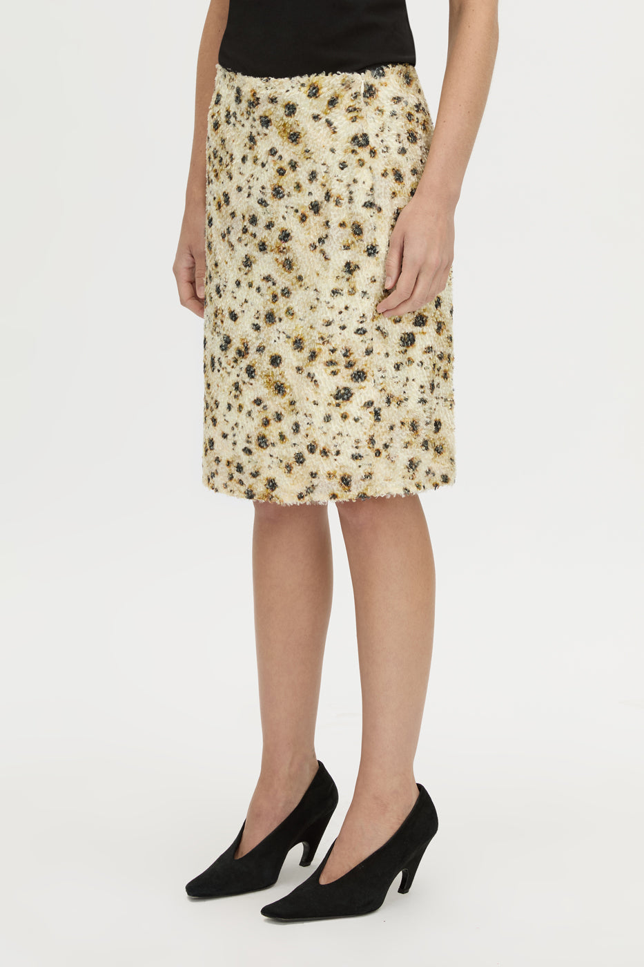 A model wearing a knee-length beige skirt with a black and brown leopard print pattern, styled with a black top and black pointed-toe heels against a plain white studio background.
