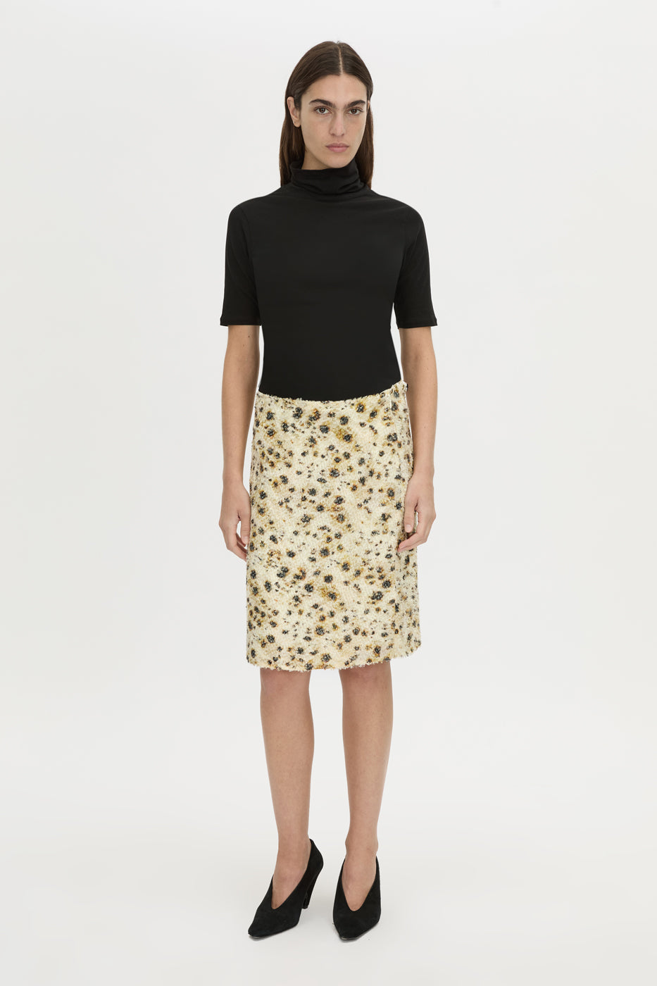 A model stands against a plain light gray studio background wearing a black short-sleeve mock-neck top and a knee-length beige skirt with a dark floral print, paired with black pointed heels.