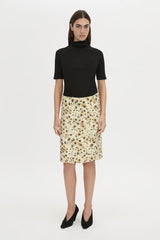 A model stands against a plain light gray studio background wearing a black short-sleeve mock-neck top and a knee-length beige skirt with a dark floral print, paired with black pointed heels.