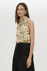 A woman modeling a sleeveless high-neck top with a beige base and black floral pattern, paired with a black skirt, against a plain light gray background.