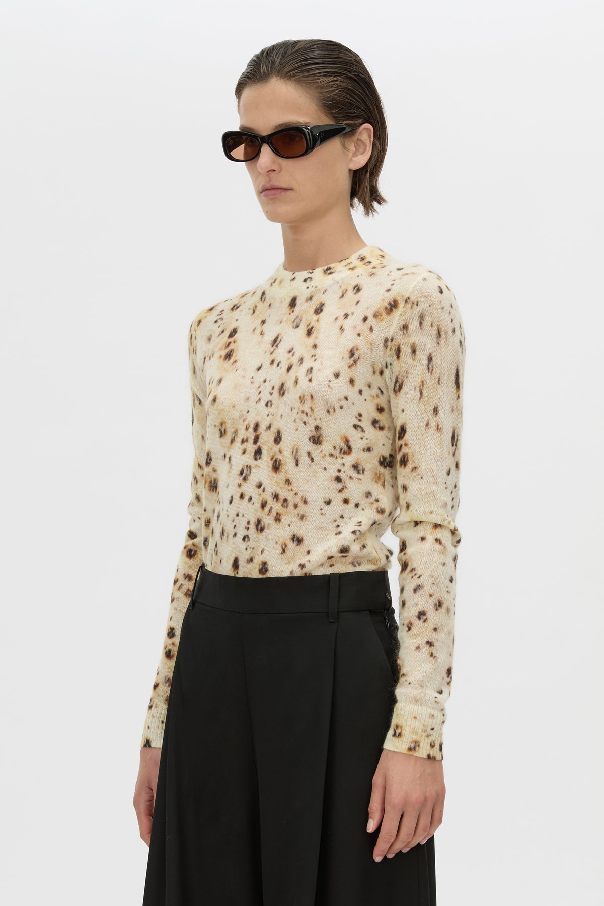 A model wearing a beige long-sleeve mock-neck top with a brown leopard print pattern, paired with black high-waisted trousers and black sunglasses, against a plain light gray background.