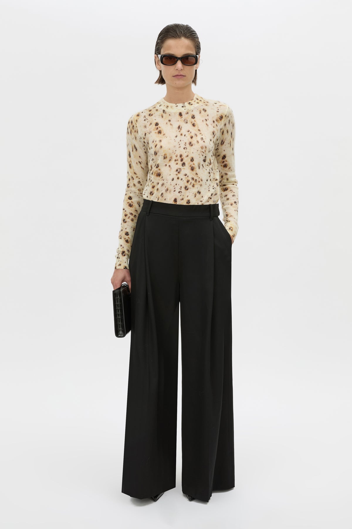 A model stands against a plain light gray studio background wearing a cream long-sleeve top with a small brown floral print, black wide-leg trousers, black sunglasses, and holding a small black clutch.