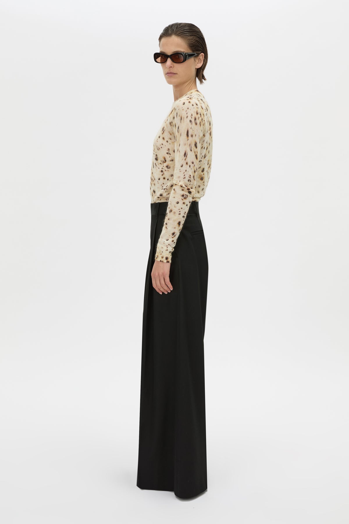 A model is shown from the side wearing a beige long-sleeve top with a brown abstract pattern and black wide-leg trousers, set against a plain white studio background.