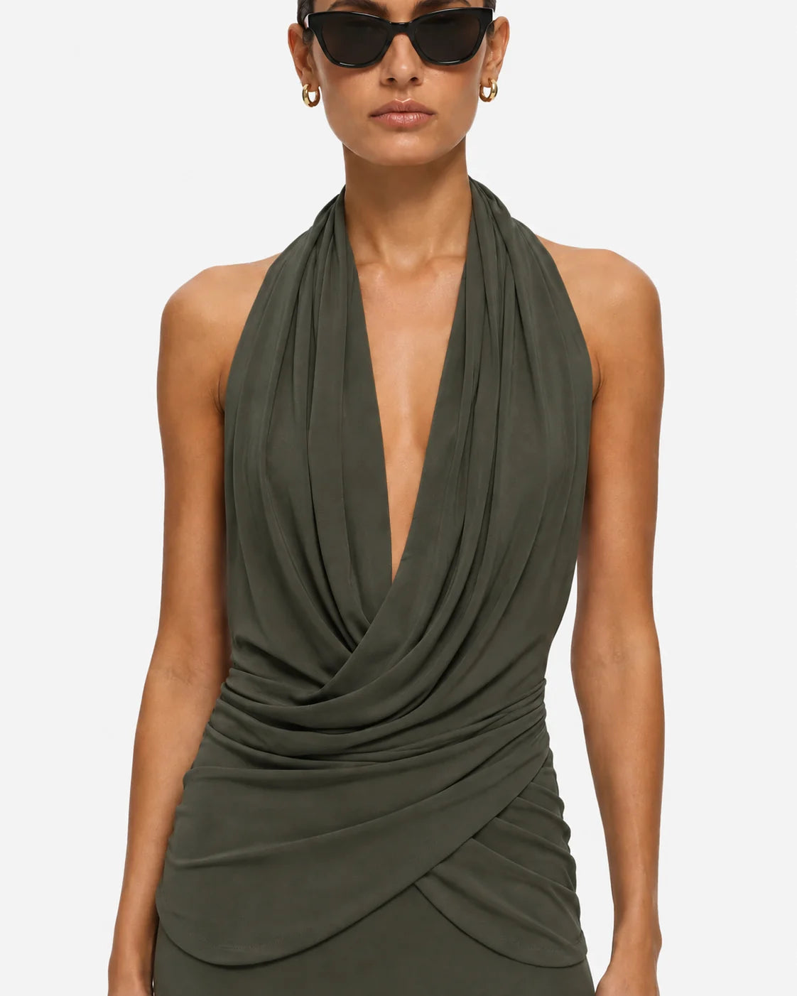 A woman wearing a dark olive green halter-neck dress with a deep V-neckline and draped, twisted fabric details at the waist, shown against a plain light gray studio background.