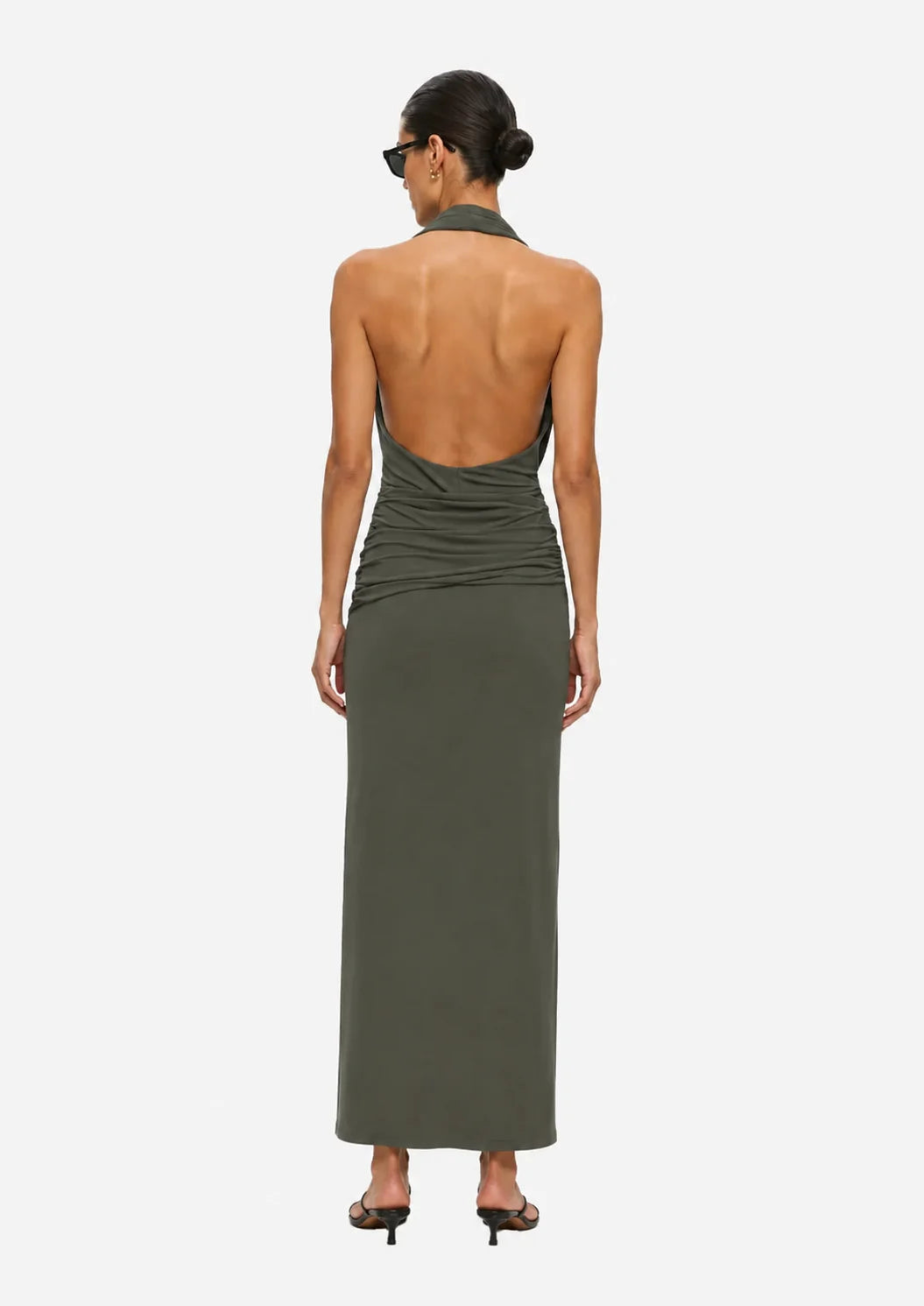 A woman viewed from the back wearing a sleeveless olive green halter maxi dress with a draped open back and gathered waist, paired with black heeled sandals against a light gray studio background.