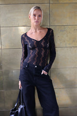 A woman stands against a metallic gold tiled wall wearing a sheer black lace long-sleeve top and dark wide-leg jeans, holding a black handbag.
