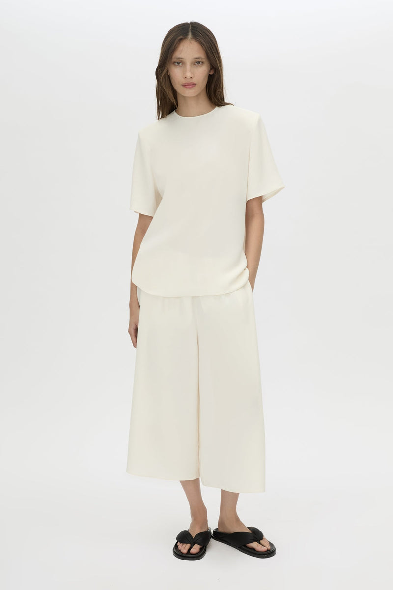 A woman modeling a matching cream-colored two-piece outfit consisting of a short-sleeve crewneck top and wide-leg cropped pants, paired with black slide sandals, against a plain white studio background.