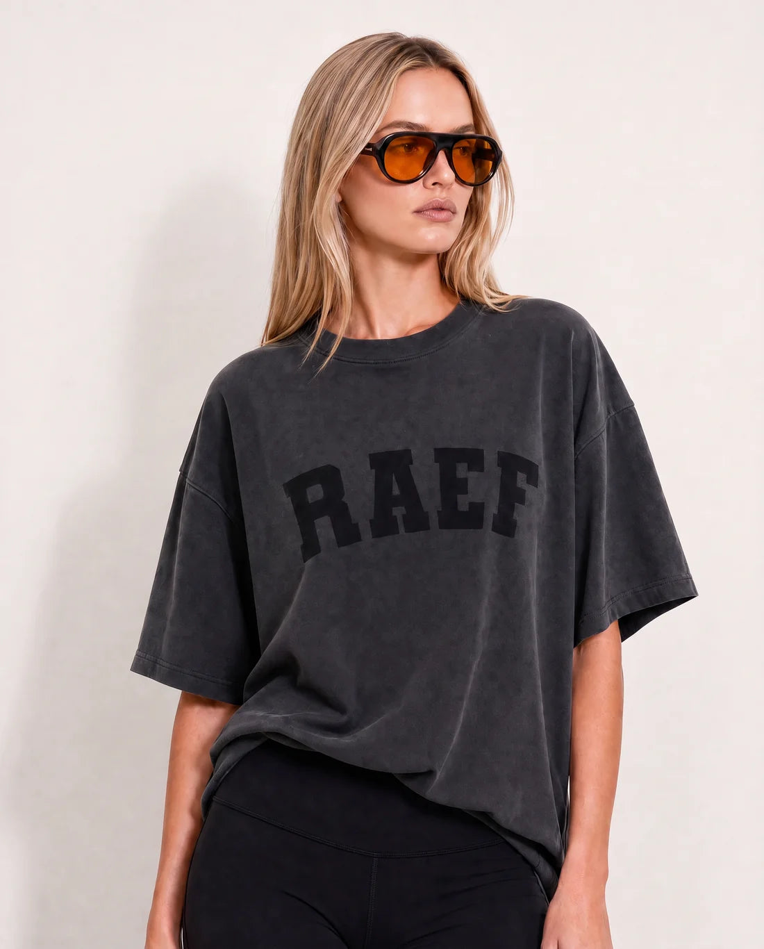 A woman modeling an oversized dark gray washed t-shirt with the word 'RAEF' printed in black arched block letters across the chest, paired with black pants and tortoiseshell sunglasses.