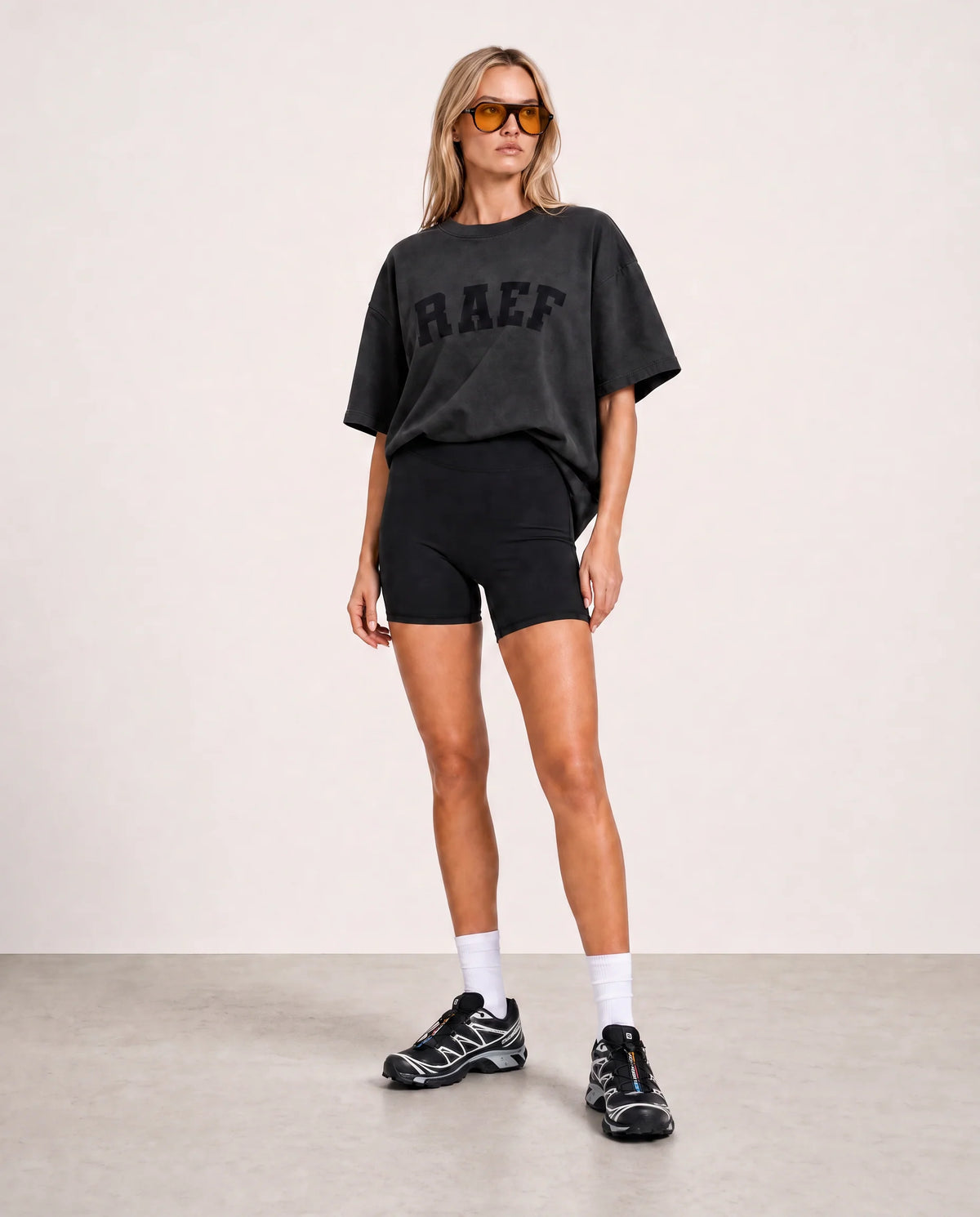 A woman modeling an oversized black graphic T-shirt with 'BAEF' printed across the chest, paired with black athletic shorts, white socks, and black sneakers, standing against a plain light beige studio background.