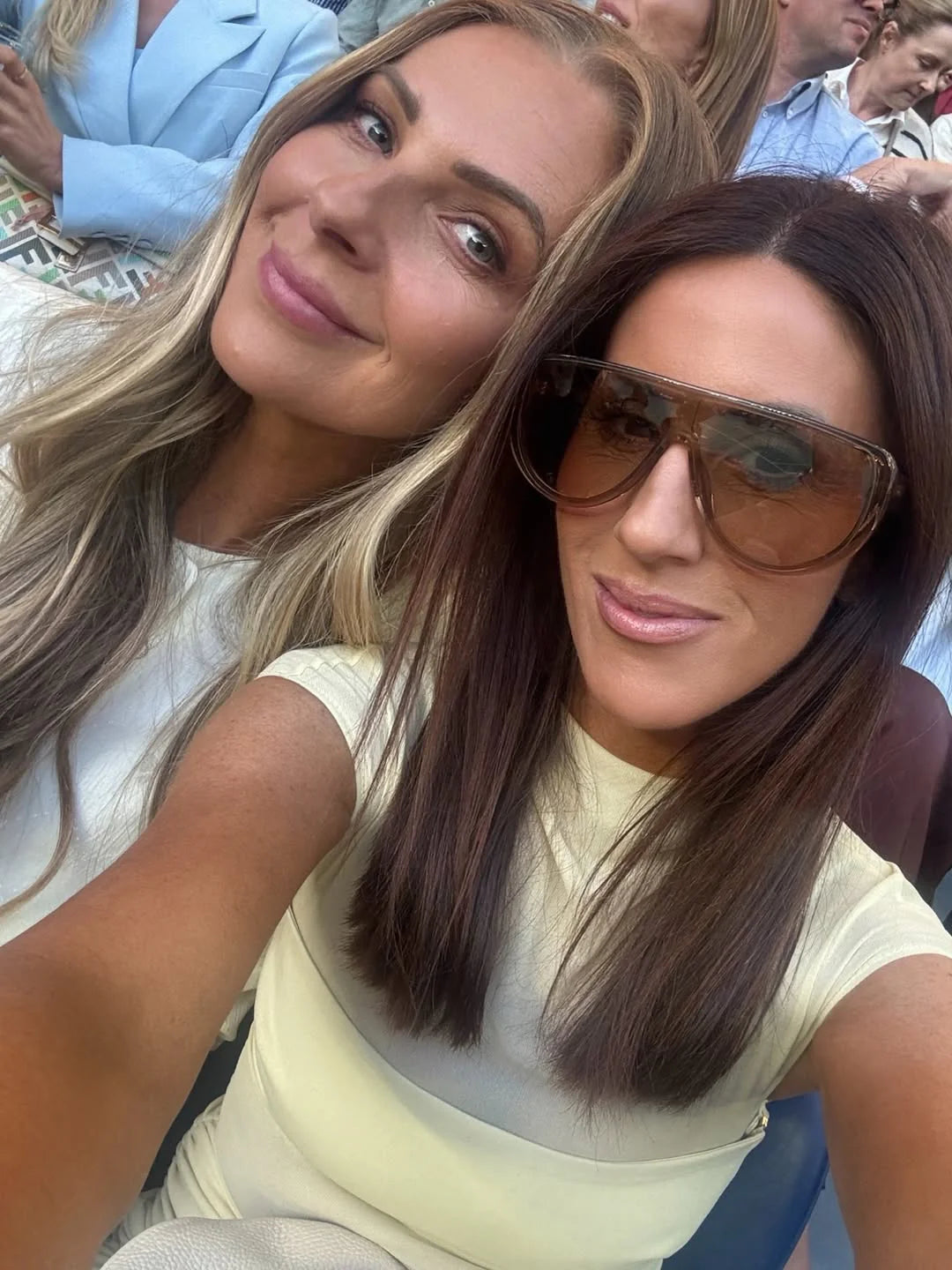 Two women taking a selfie together, one wearing sunglasses, with a crowd in the background.