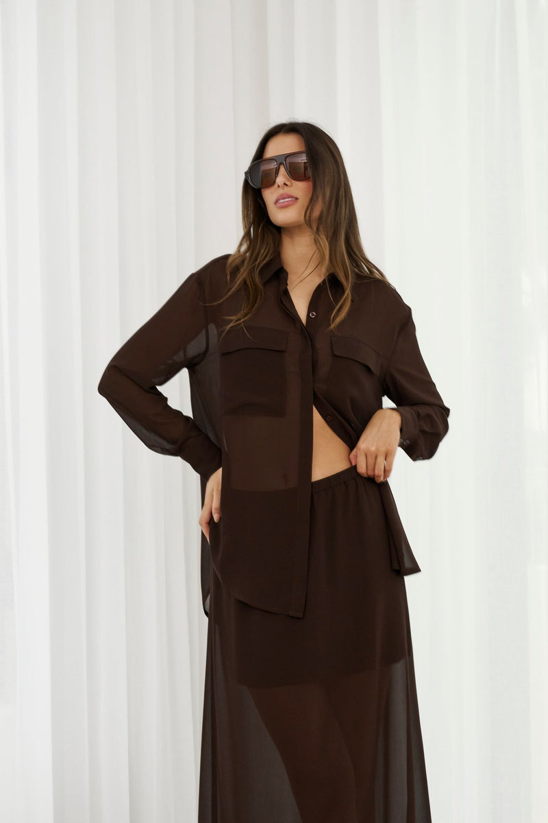 Woman wearing a brown outfit with sunglasses against a white curtain background