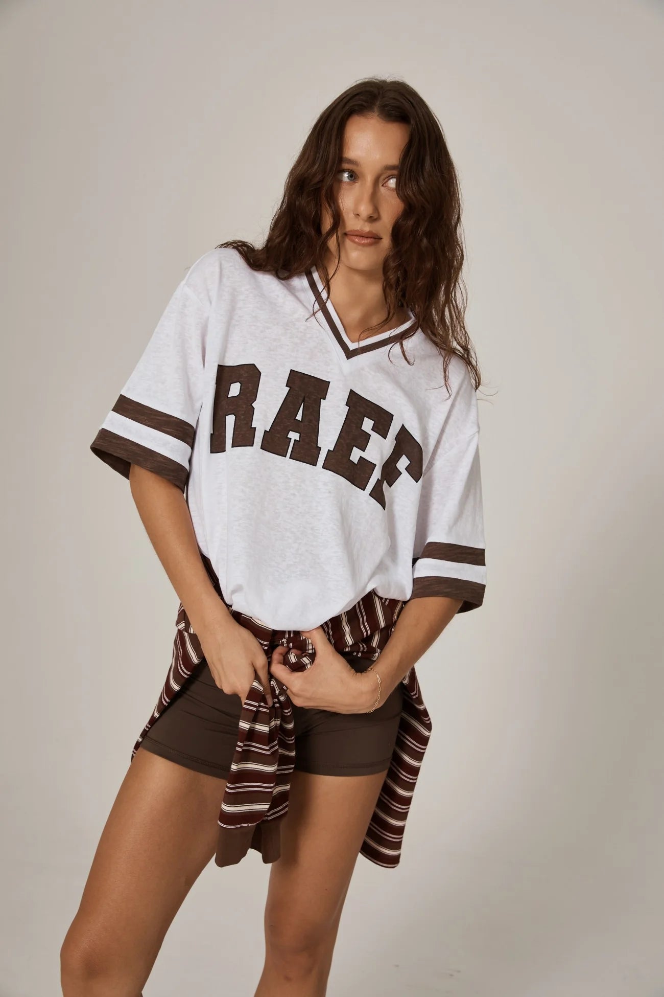 Woman standing in a cool girl white oversized tee with brown graphics and a stripe sweater around her waist