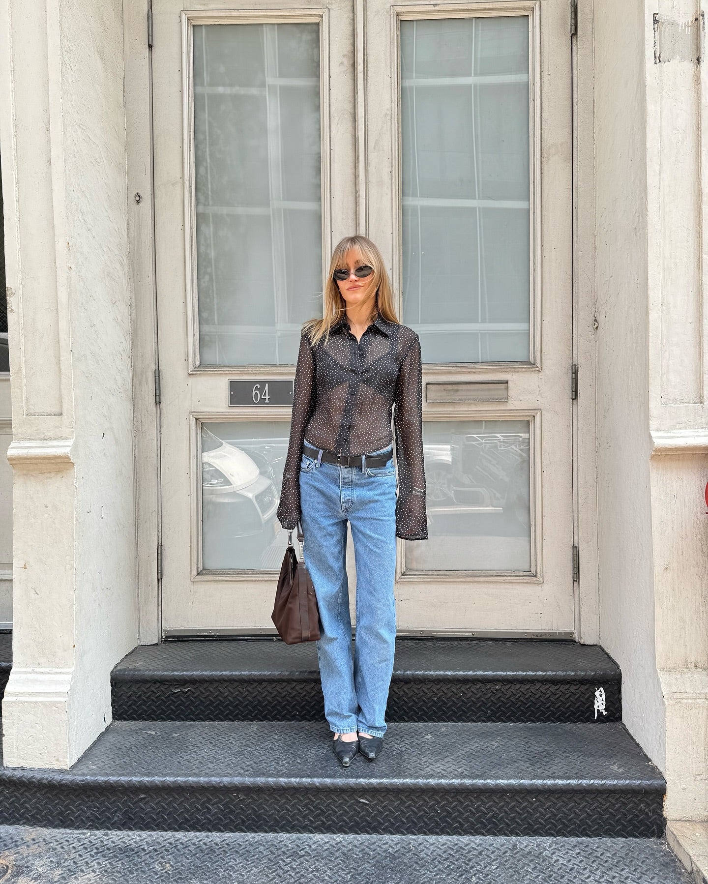 A woman stands on the black diamond-plate steps of a building entrance, wearing a sheer brown long-sleeve button-up shirt, light blue straight-leg jeans, a black belt, and black shoes, while holding a brown leather handbag.