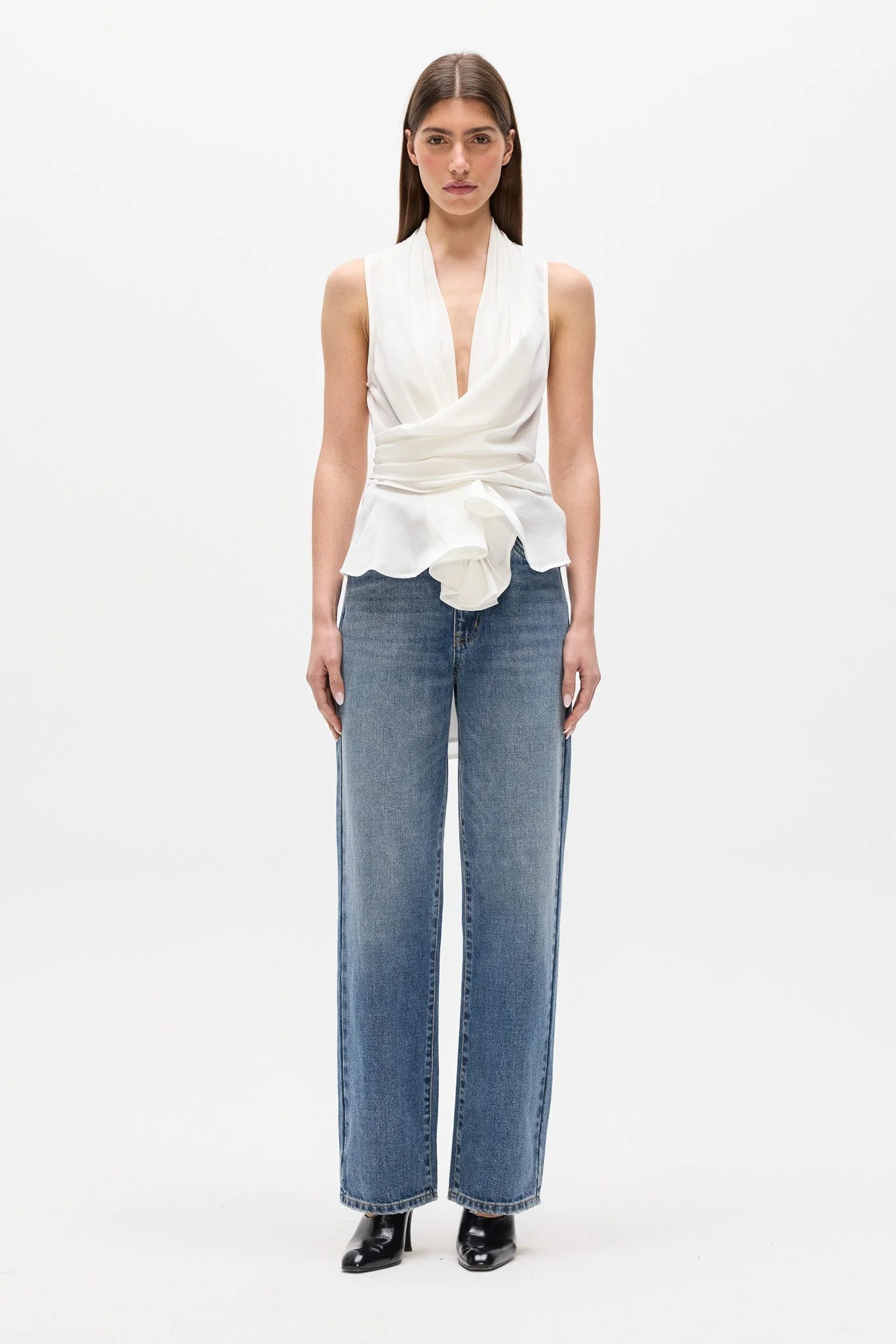 A model stands against a plain white studio background wearing a sleeveless white wrap-style top with a deep V-neck and gathered waist, paired with wide-leg blue jeans and black pointed shoes.