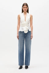 A model stands against a plain white studio background wearing a sleeveless white wrap-style top with a deep V-neck and gathered waist, paired with wide-leg blue jeans and black pointed shoes.