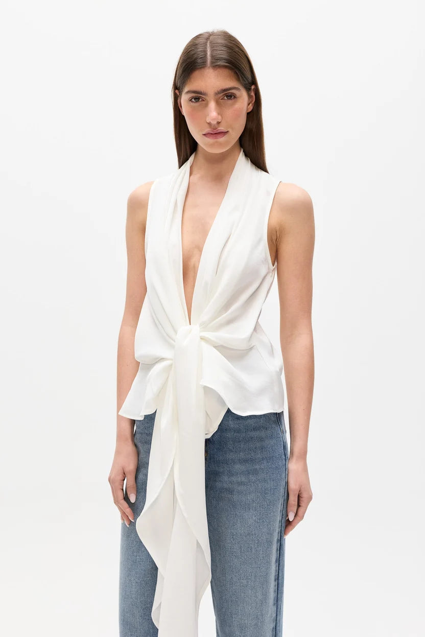 A woman modeling a sleeveless white wrap-style top with a deep V-neck and front tie detail, paired with blue jeans, against a plain white studio background.