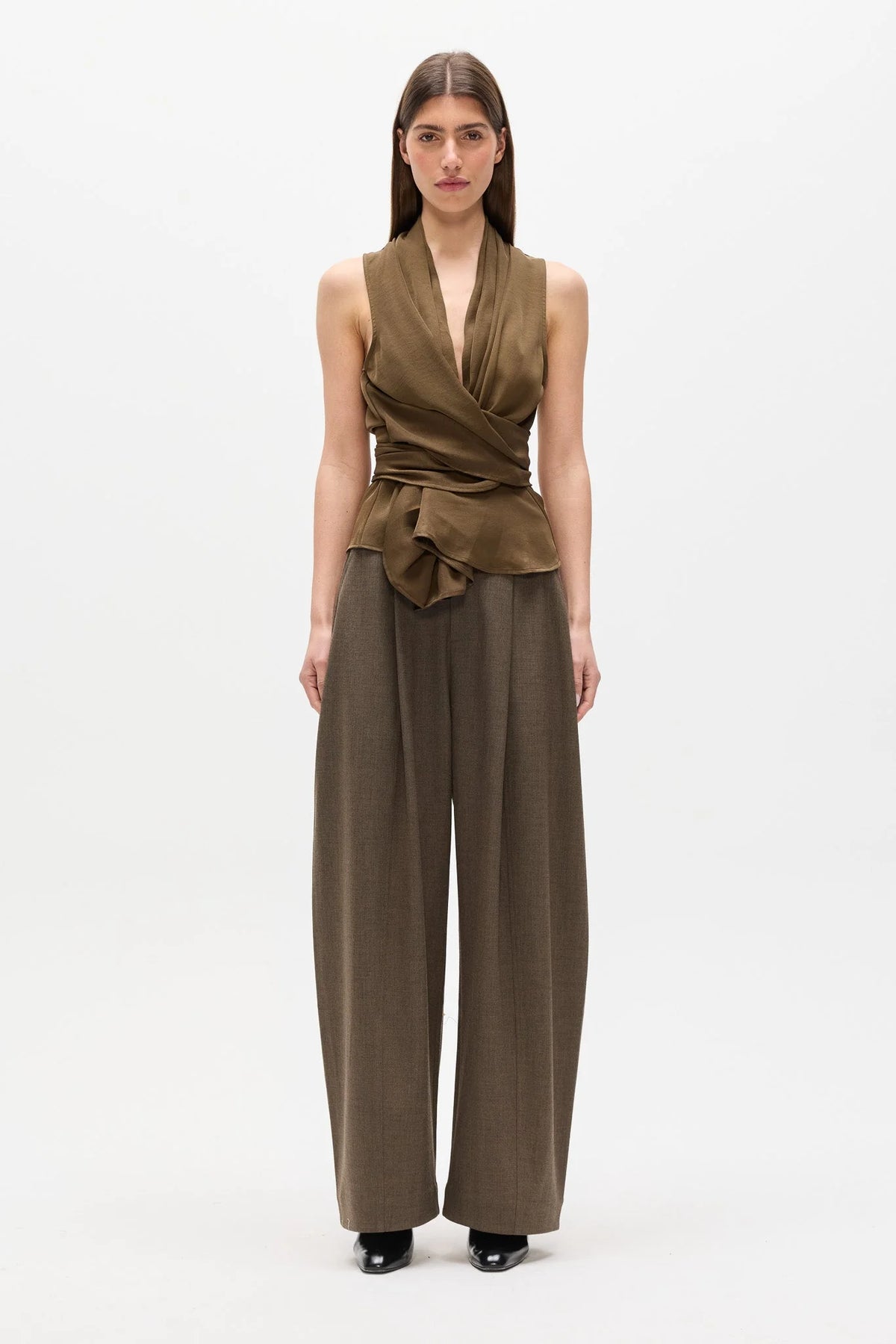 A model stands against a plain white studio background wearing a sleeveless wrap-style top in olive brown with matching wide-leg trousers and black shoes.