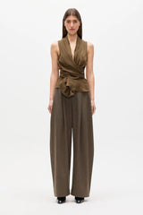A model stands against a plain white studio background wearing a sleeveless wrap-style top in olive brown with matching wide-leg trousers and black shoes.