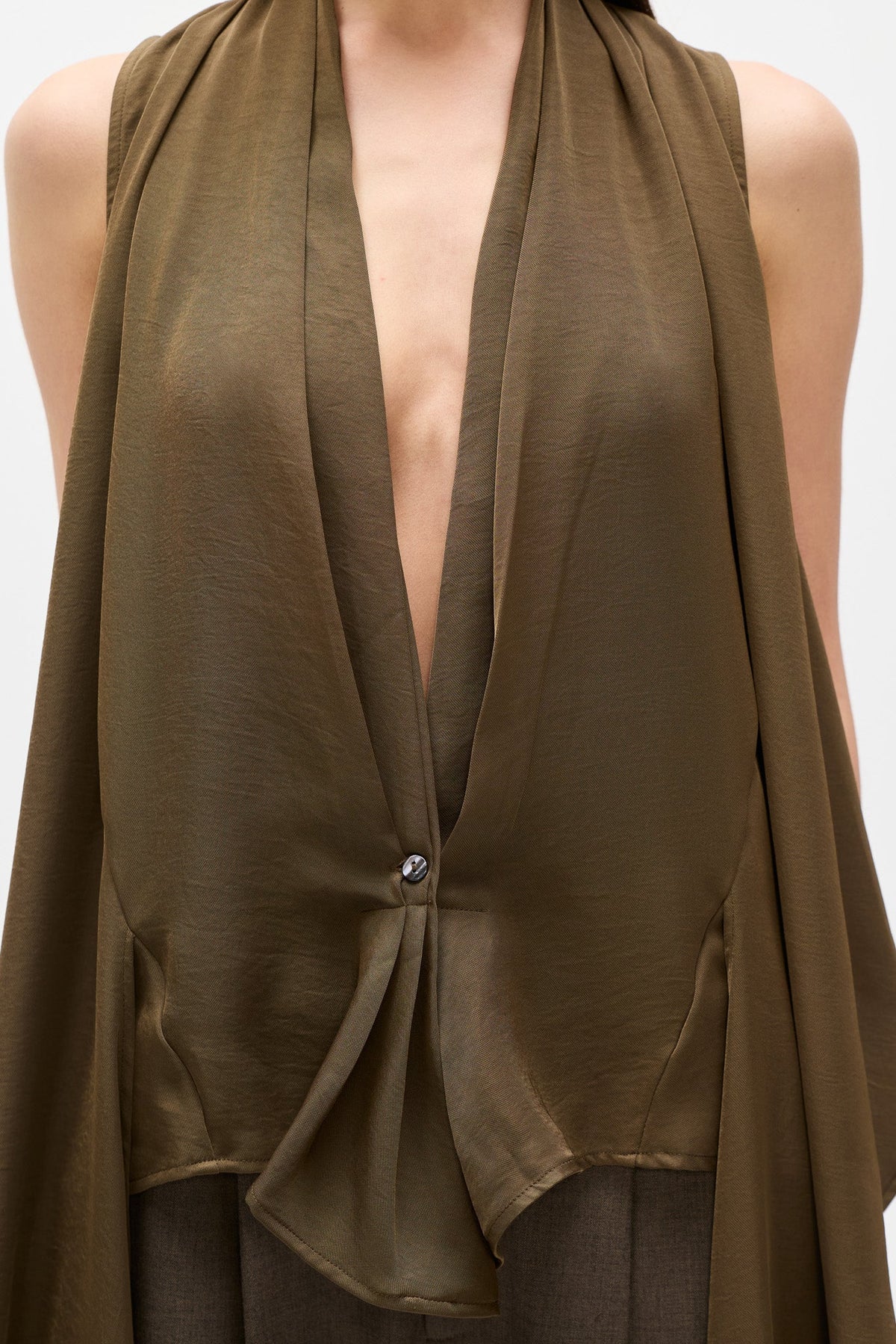 A close-up of a model wearing a sleeveless, deep V-neck draped top in olive brown. The top features a single button closure near the center and is styled over dark trousers against a plain light background.