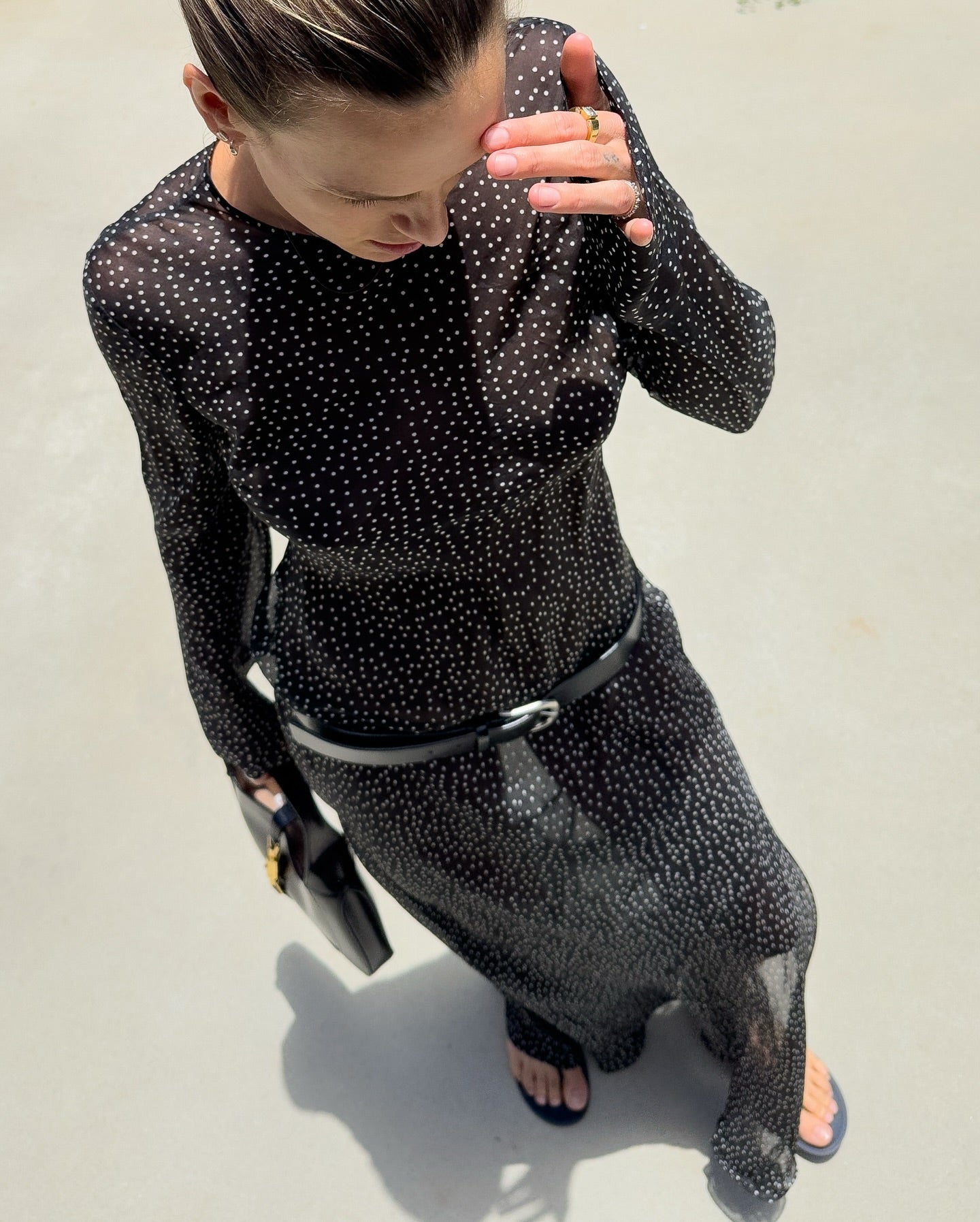 A woman wearing a sheer black long-sleeve maxi dress with a small white polka dot pattern, styled with a black belt and black sandals, photographed from a high-angle outdoor perspective.