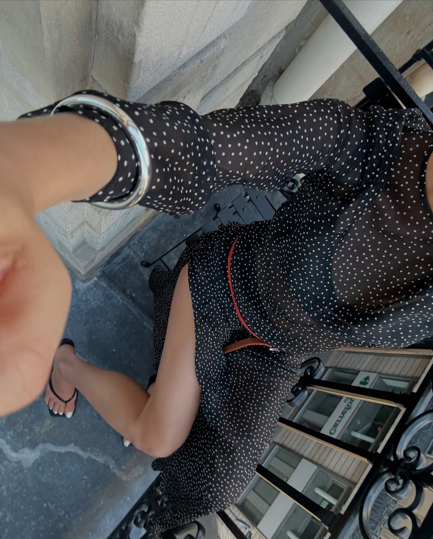A person wearing a sheer black long-sleeve dress with a white polka dot pattern, taking a selfie on outdoor stone steps.