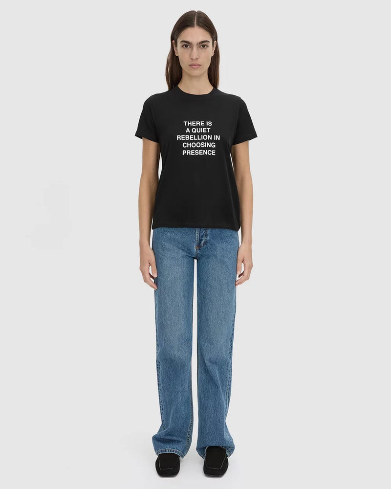 A model stands against a light gray studio background wearing a black graphic T-shirt with white text, blue jeans, and black shoes. The shirt reads, 'THERE IS A QUIET REBELLION IN CHOOSING PRESENCE.