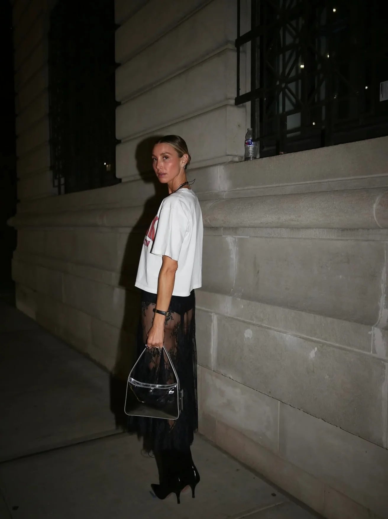 Person standing on steps holding a clear handbag at night in a white Tshirt at fashion week
