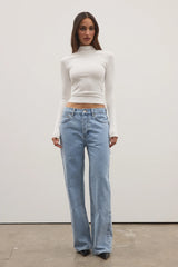 Woman wearing a white turtleneck and light blue jeans against a plain background