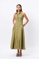 Woman standing in an olive dress. Crafted from premium Australian cotton, this sleeveless midi features a structured collared neckline, racerback detail, and a fitted bodice that flows into a semi-flared A-line skirt. Stitch detailing at the fixed waist enhances the silhouette, while functional side pockets add everyday ease. Designed for the modern minimalist, this effortless style transitions seamlessly from day to evening.