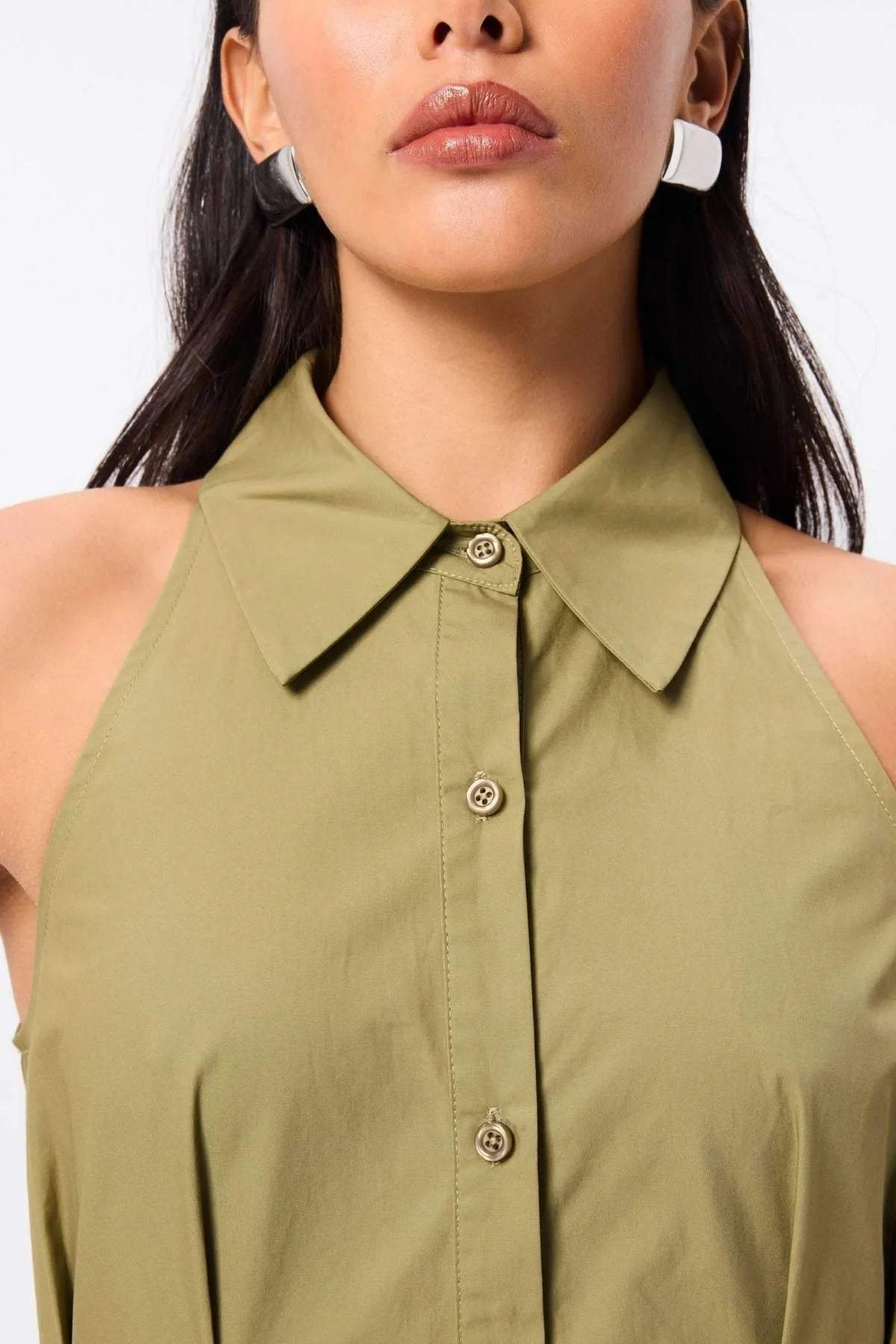 Woman standing in an olive dress. Crafted from premium Australian cotton, this sleeveless midi features a structured collared neckline, racerback detail, and a fitted bodice that flows into a semi-flared A-line skirt. Stitch detailing at the fixed waist enhances the silhouette, while functional side pockets add everyday ease. Designed for the modern minimalist, this effortless style transitions seamlessly from day to evening.