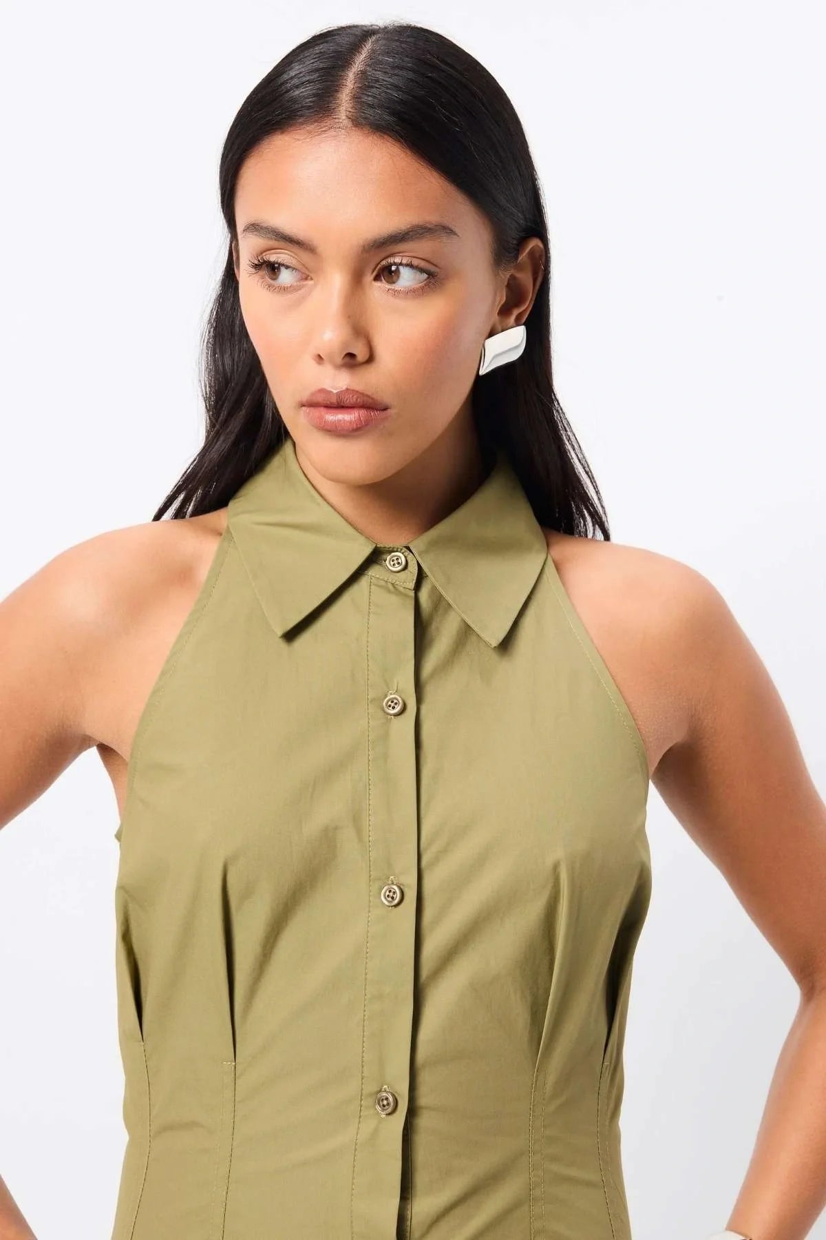 Woman standing in an olive dress. Crafted from premium Australian cotton, this sleeveless midi features a structured collared neckline, racerback detail, and a fitted bodice that flows into a semi-flared A-line skirt. Stitch detailing at the fixed waist enhances the silhouette, while functional side pockets add everyday ease. Designed for the modern minimalist, this effortless style transitions seamlessly from day to evening.