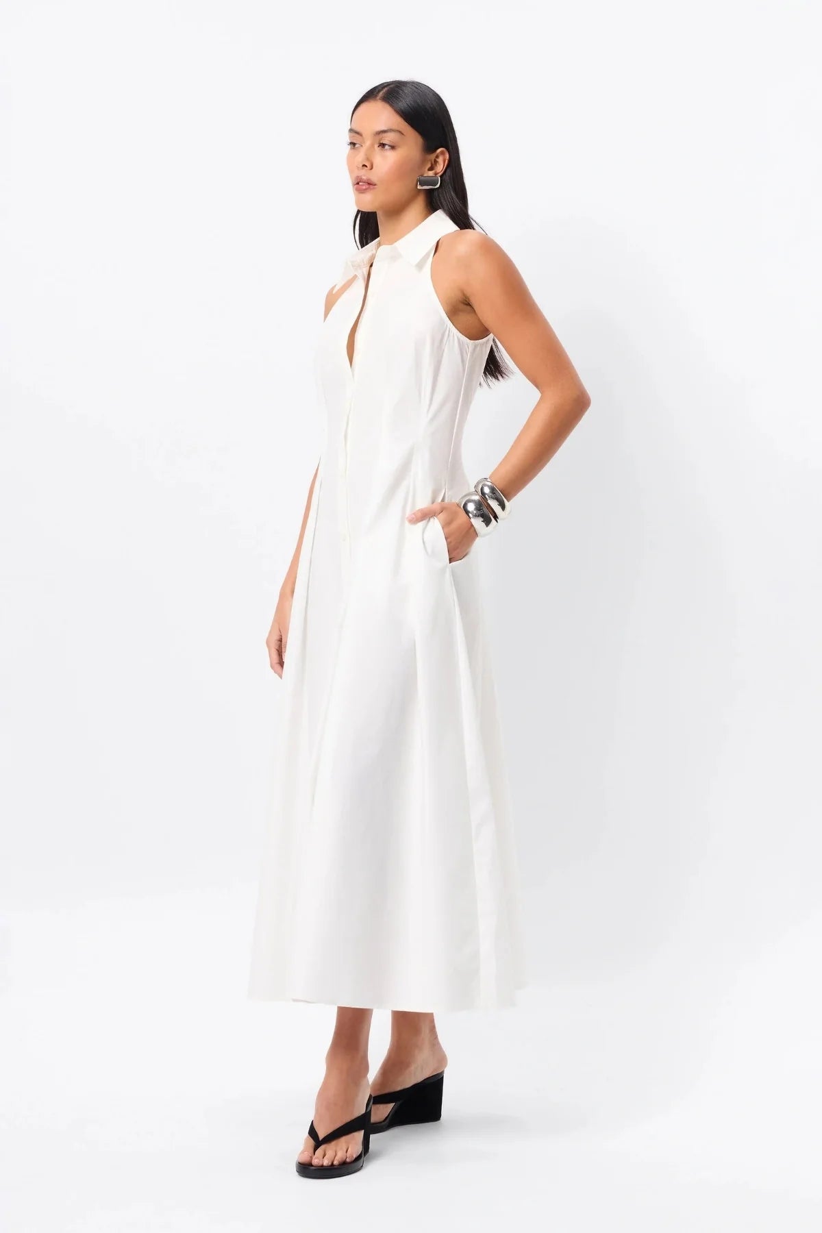 A woman standing in a white cotton day dress with pleat detail— a refined take on the classic shirt dress with timeless appeal.

Crafted from premium Australian cotton, this sleeveless midi features a structured collared neckline, racerback detail, and a fitted bodice that flows into a semi-flared A-line skirt. Stitch detailing at the fixed waist enhances the silhouette, while functional side pockets add everyday ease. Designed for the modern minimalist, this effortless style transitions seamlessly from day