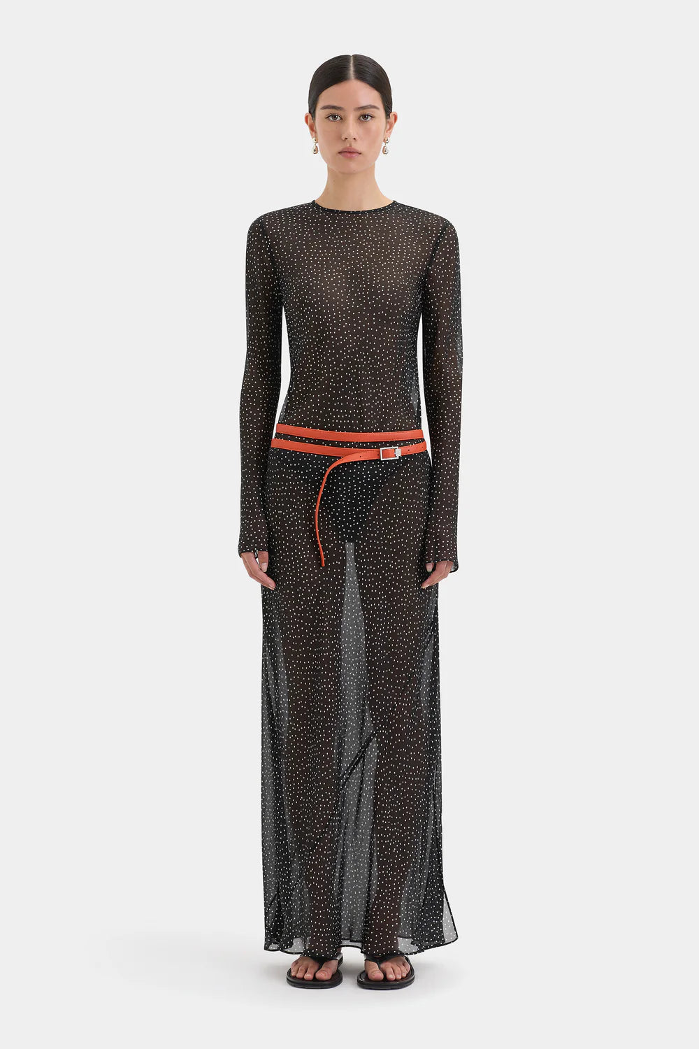 A model stands against a light gray studio background wearing a sheer black long-sleeve maxi dress with a small repeating pattern, a bright orange-red waist belt, and black sandals.