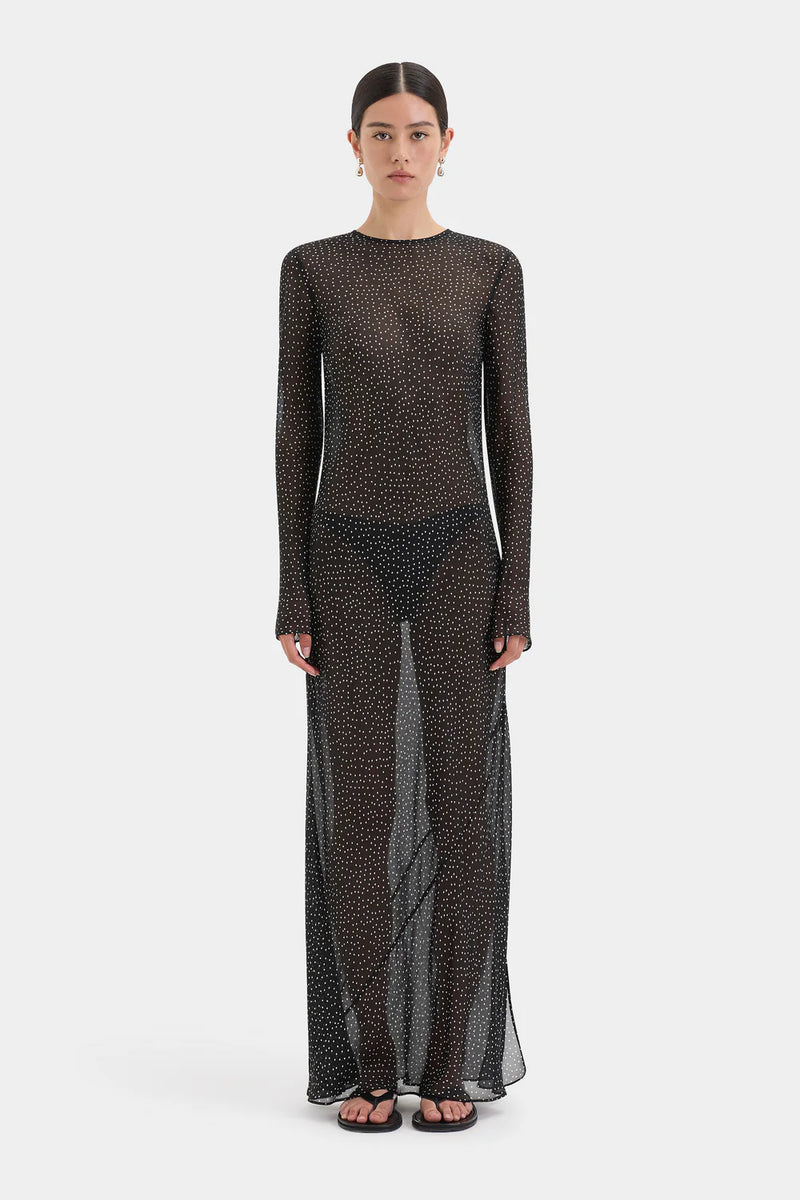 A model wearing a sheer, long-sleeved maxi dress with a dense black and brown abstract pattern, standing against a light gray studio background.