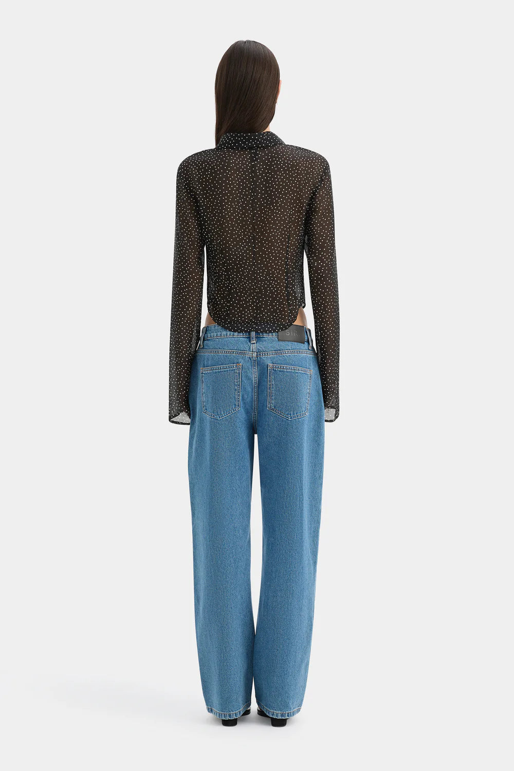 A model shown from the back wearing a sheer black long-sleeve top with a small brown pattern, paired with high-waisted, wide-leg blue jeans, against a light gray studio background.