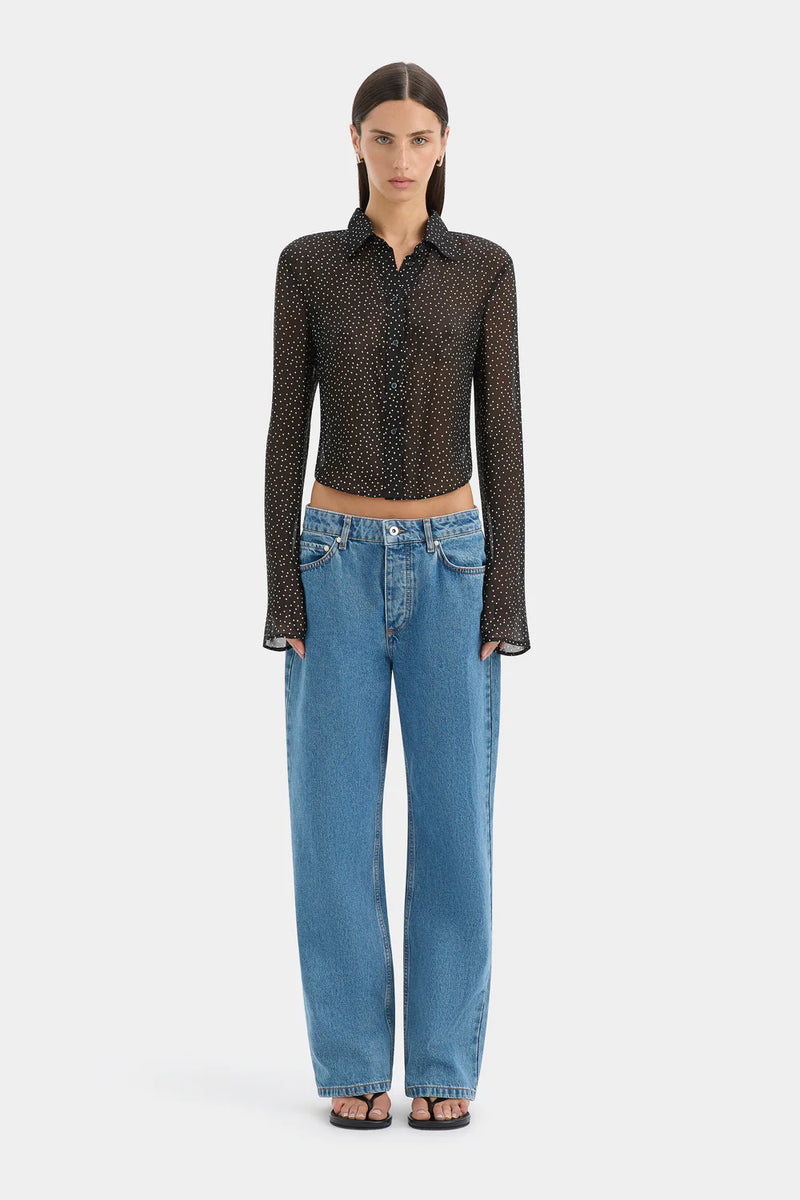 A model stands against a light gray studio background wearing a sheer black long-sleeve button-up shirt with a small repeating pattern, paired with wide-leg blue jeans and black sandals.