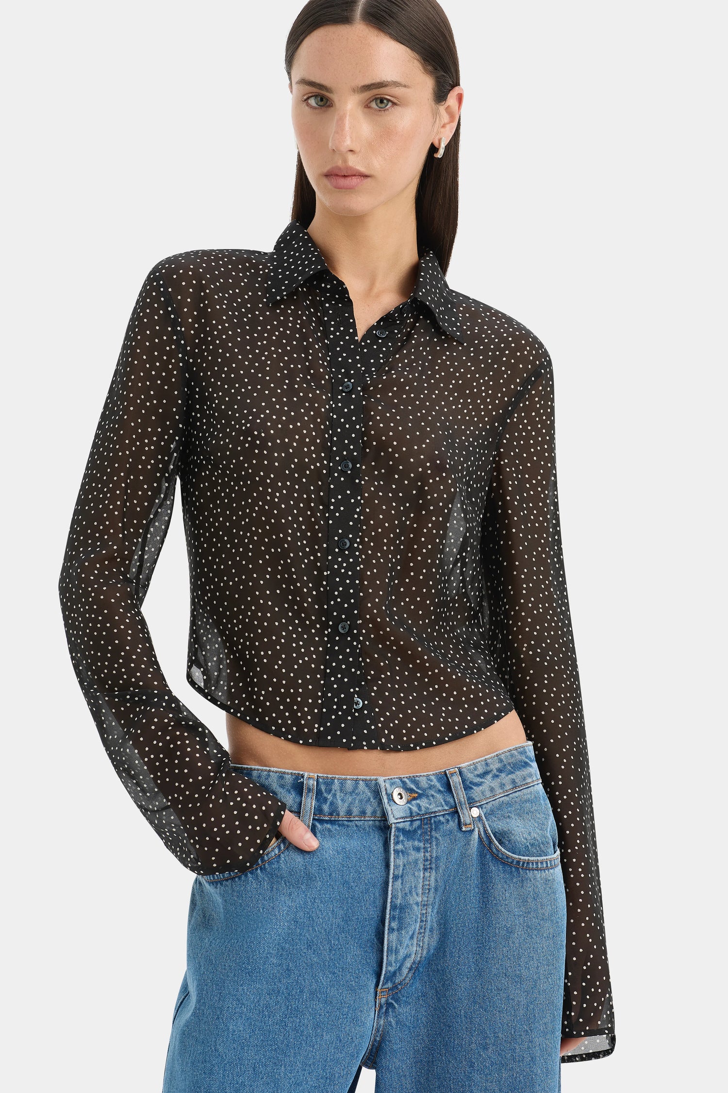 A woman modeling a sheer black long-sleeve button-up shirt with a white polka-dot pattern, paired with blue jeans, against a light gray studio background.