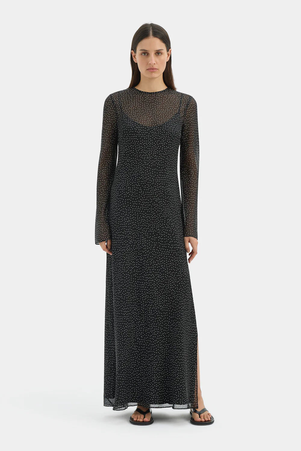 A woman modeling a long-sleeve black maxi dress with a subtle white micro-pattern and a sheer mesh yoke, standing against a light gray studio background.
