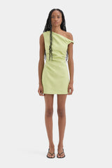 A model wearing a light lime green one-shoulder bodycon mini dress with silver chain accents on the side, paired with black strappy sandals, against a plain light gray background.