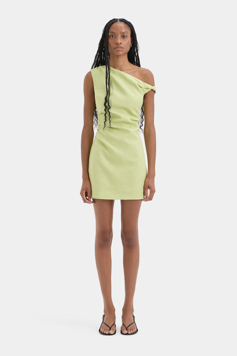 A model wearing a light lime green one-shoulder bodycon mini dress with silver chain accents on the side, paired with black strappy sandals, against a plain light gray background.