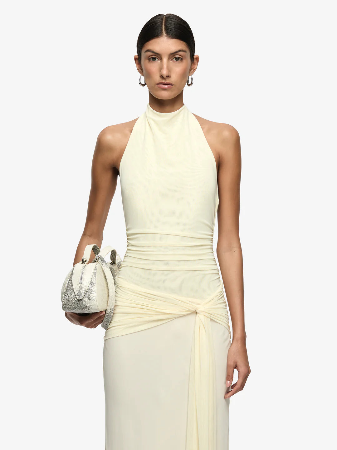 Woman wearing a light-colored halter neck dress holding a matching clutch against a white background