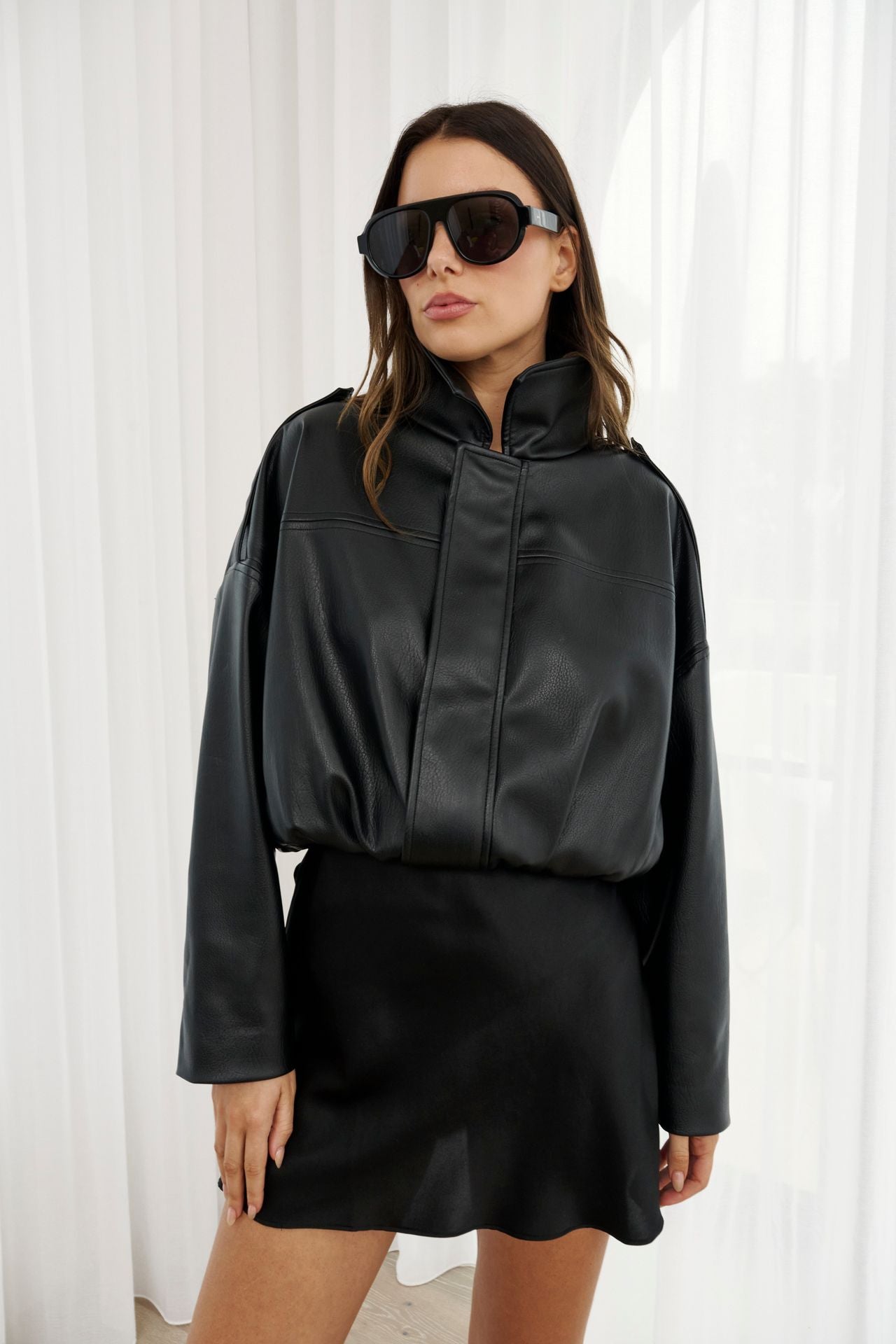 Woman wearing a black leather outfit with sunglasses against a white curtain background