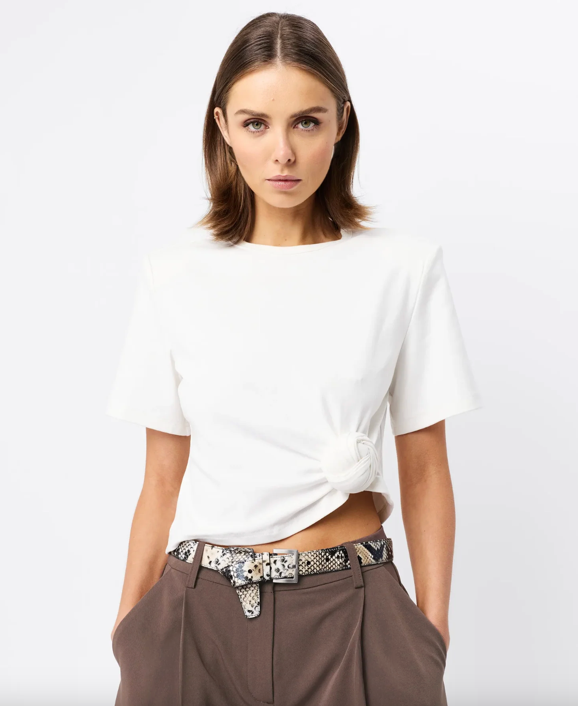 Woman wearing a white t-shirt and brown pants with a patterned belt on a white background